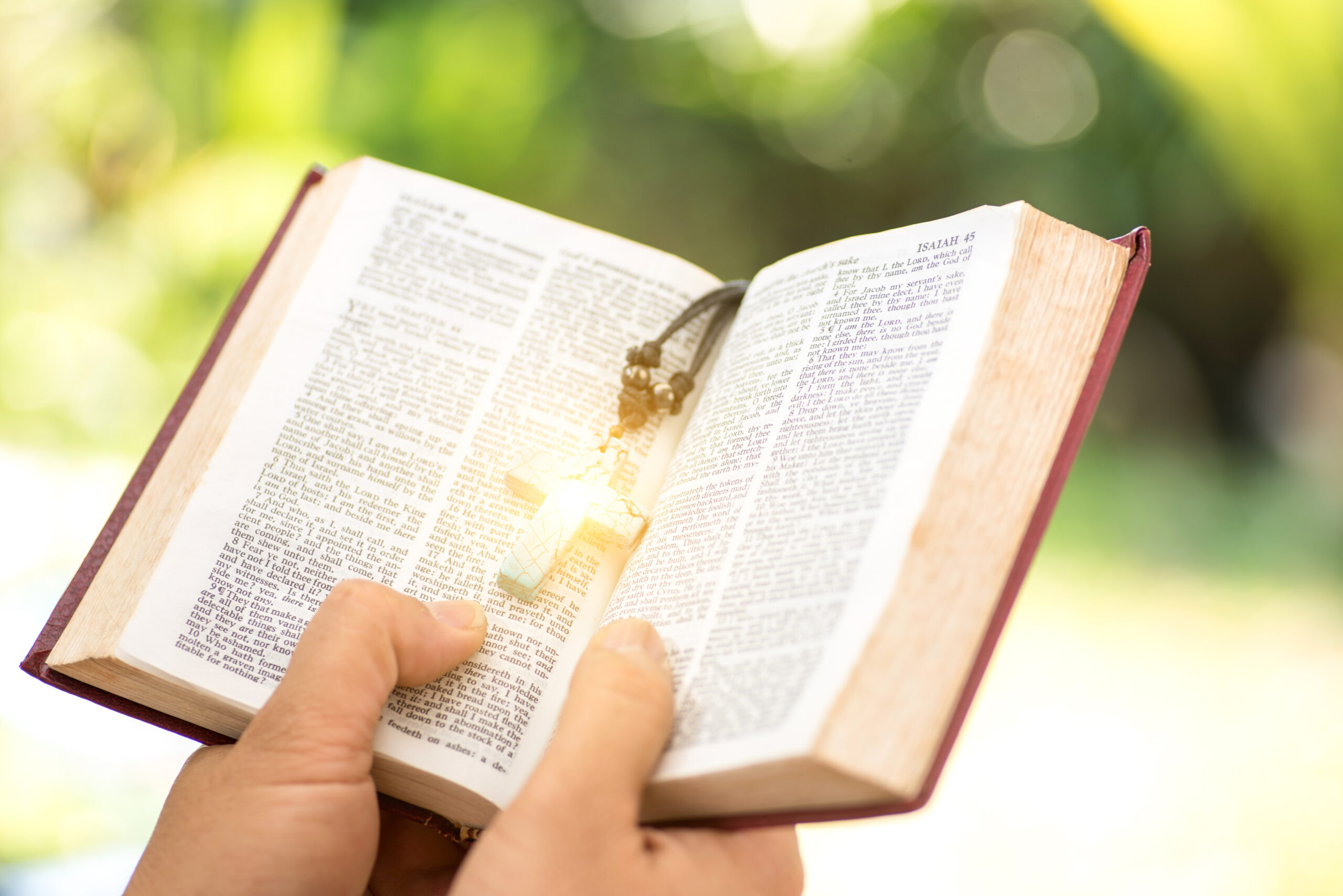 hands holding an opened Bible with a cross book mark with the sun shinning on the book mark